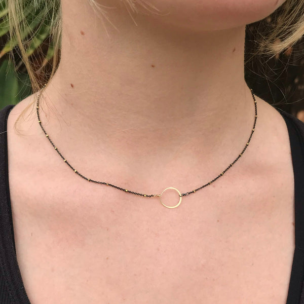 dainty handmade necklace with gold-filled tiny circle on oxidized silver, black chain with tiny gold beads on gal wearing black shirt in front of palm fronds